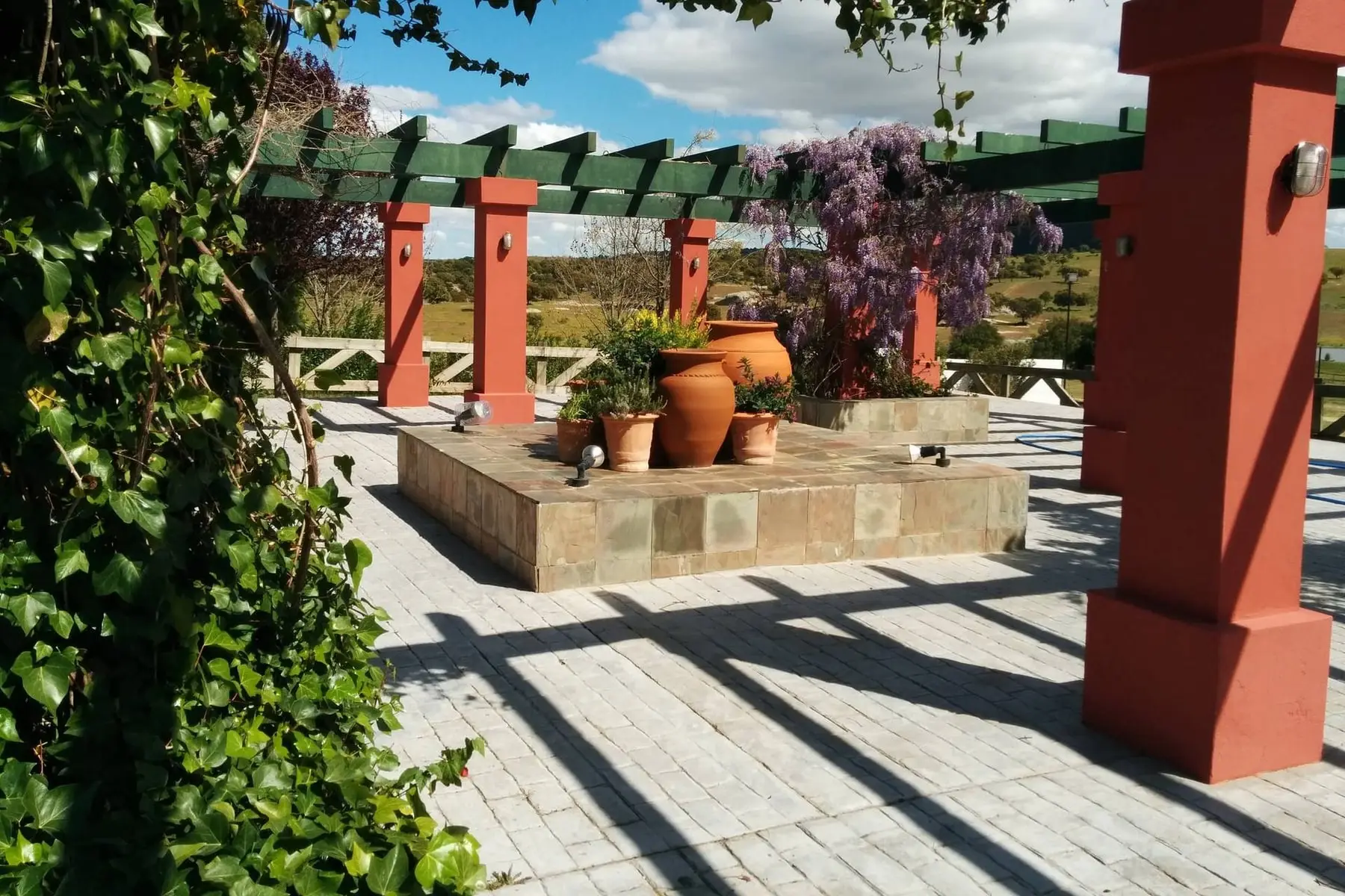 La pérgola de columnas rojas, con glicinia, una tinaja de barro y un banco de pizarra